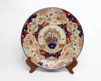 Large wall plate, Japan, Arita, Meiji period (1868-1912), late 19th century, porcelain, Imari style decorated in underglaze blue, iron red, green and gold, in the centre a vase with blooming peonies and willow branches on a terrace,...
