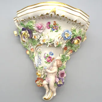MANUFACTURE DE PORCELAINE SAXONNE DRESDEN POTSCHAPPEL. CONSOLE MURALE AVEC PUTTO, DÉCOR FLORAL EN RELIEF ET DÉTAILS DORÉS. DÉBUT DU 20E SIÈCLE.
