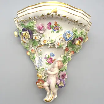 MANUFACTURE DE PORCELAINE SAXONNE DRESDEN POTSCHAPPEL. CONSOLE MURALE AVEC PUTTO, DÉCOR FLORAL EN RELIEF ET DÉTAILS DORÉS. DÉBUT DU 20E SIÈCLE.